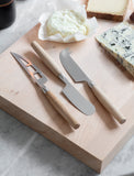 Two cheese knives on a wooden cutting board with cheese and bread in the background.