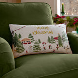 A rectangular Christmas throw pillow with a winter village scene and gold 'Merry Christmas' script, resting on an olive green armchair in a cozy indoor setting.