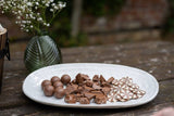 A white oval plate filled with assorted chocolates, including whole milk chocolate chunks, dark chocolate balls, and decorated milk chocolate flowers, set on a rustic wooden table with a blurred green glass vase of baby's breath in the background.
