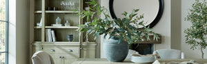 A tastefully decorated dining or living room featuring a wooden cabinet, a large ceramic vase with lush greenery on a table, a round wall mirror, and neutral-toned seating, showcasing KHEE's interior style.