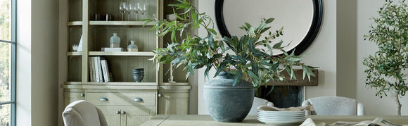 A tastefully decorated dining or living room featuring a wooden cabinet, a large ceramic vase with lush greenery on a table, a round wall mirror, and neutral-toned seating, showcasing KHEE's interior style.
