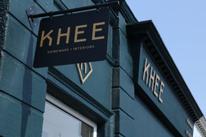 xterior view of the KHEE Homeware & Interiors storefront, featuring the black and gold KHEE brand sign prominently displayed on a dark teal building facade under a clear blue sky.